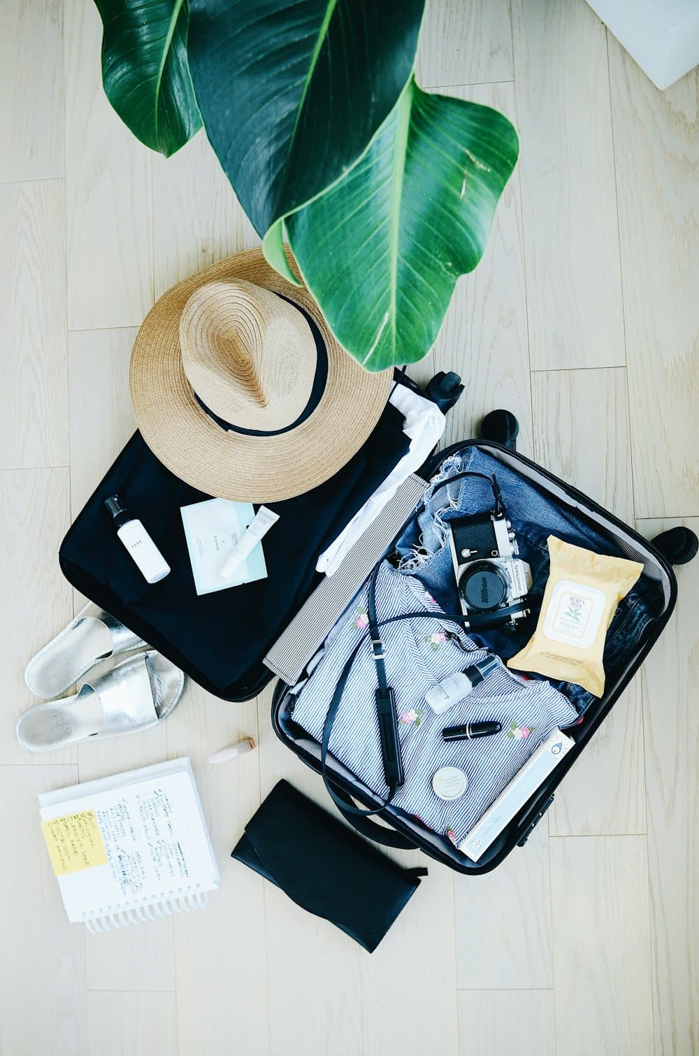 An open suitcase with a straw hat, camera and travel items on a wooden floor
