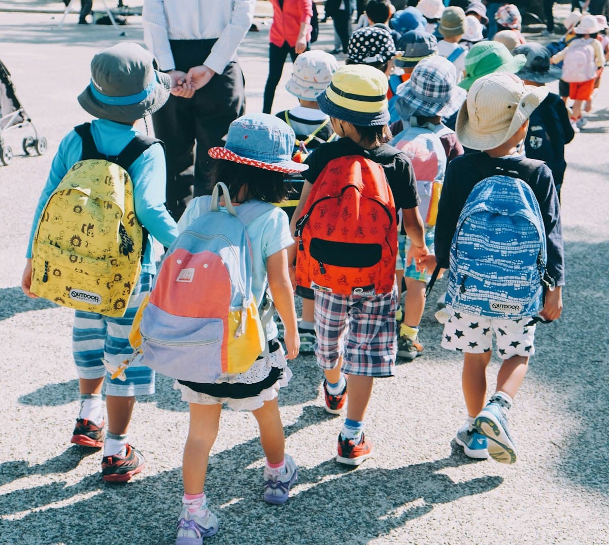A group of small children in sun hats walking to school with colourful backpacks